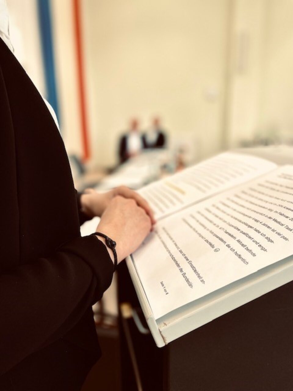 Person hält ein Manuskript an einem Pult, im Hintergrund unscharfe sitzende Personen.