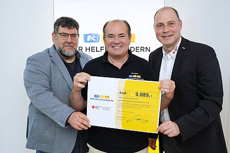 Drei Männer halten einen Scheck über 9.089 Euro für den DRK-Landesverband, mit "Wir helfen Kindern"-Logo im Hintergrund.