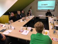 Personen in einem Sitzungsraum, Projektpräsentation auf Leinwand, Tische mit Getränken und Notizmaterial.