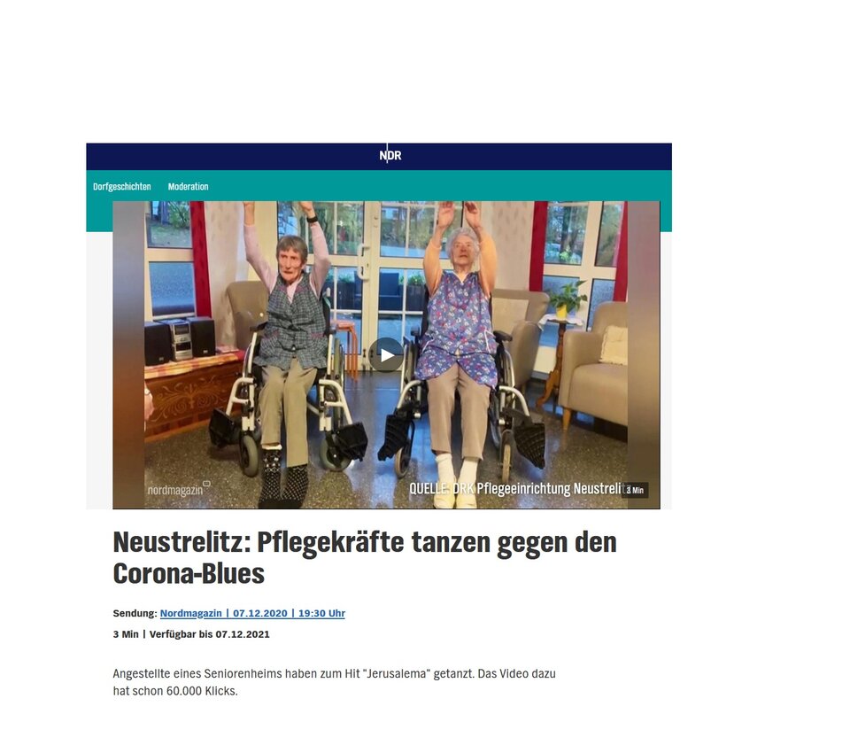 Zwei lachende Personen tanzen im Rollstuhl in einem Raum mit Fenstern.