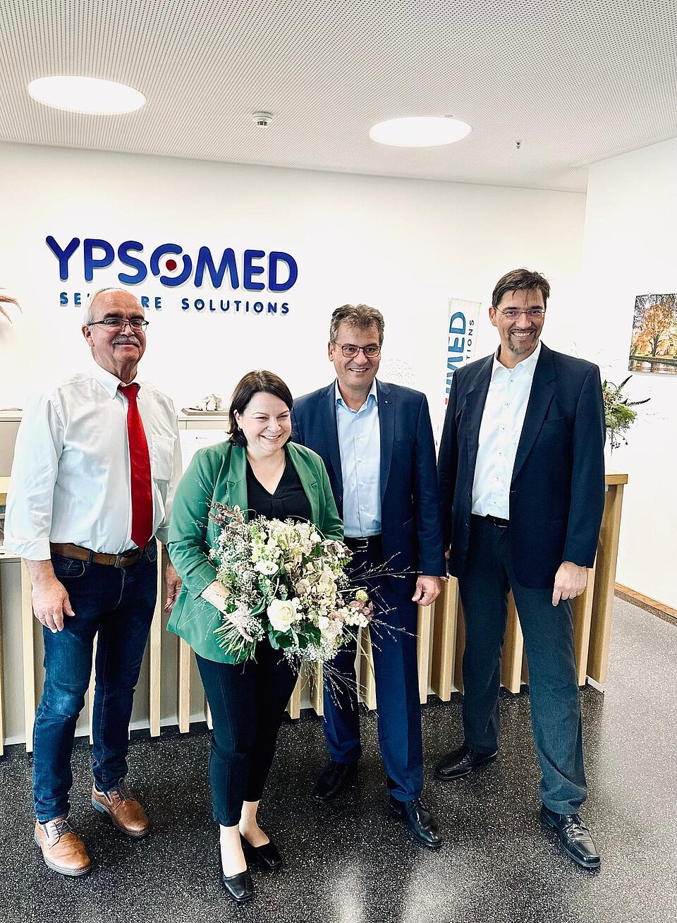 Vier Personen stehen lächelnd in einem Büro vor einem Ypsomed-Logo, eine Frau hält einen Blumenstrauß.
