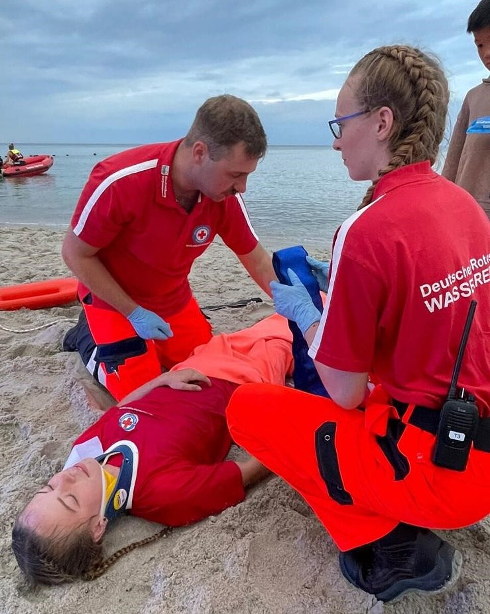 Rettungskräfte versorgen verletzte Person am Strand. Eine trägt einen Halskragen. Boot im Hintergrund.