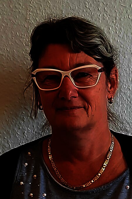 Person mit Brille vor einer hellen Wand, trägt eine Kette und ein graues Oberteil mit Perlendetails.