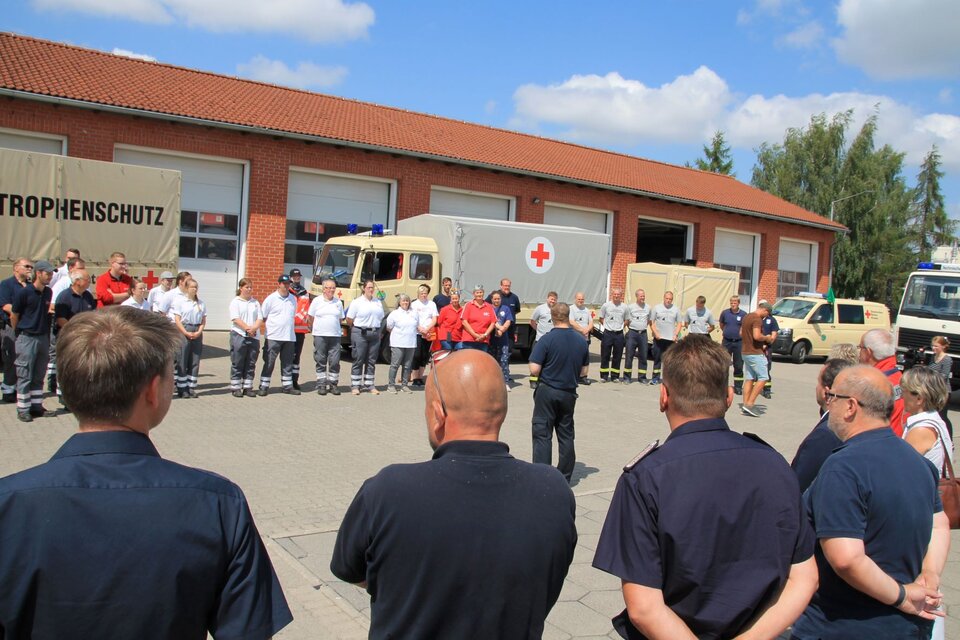 Gruppe von Einsatzkräften versammelt sich vor Fahrzeugen mit Katastrophenschutz-Aufschrift vor einer Backsteinhalle.