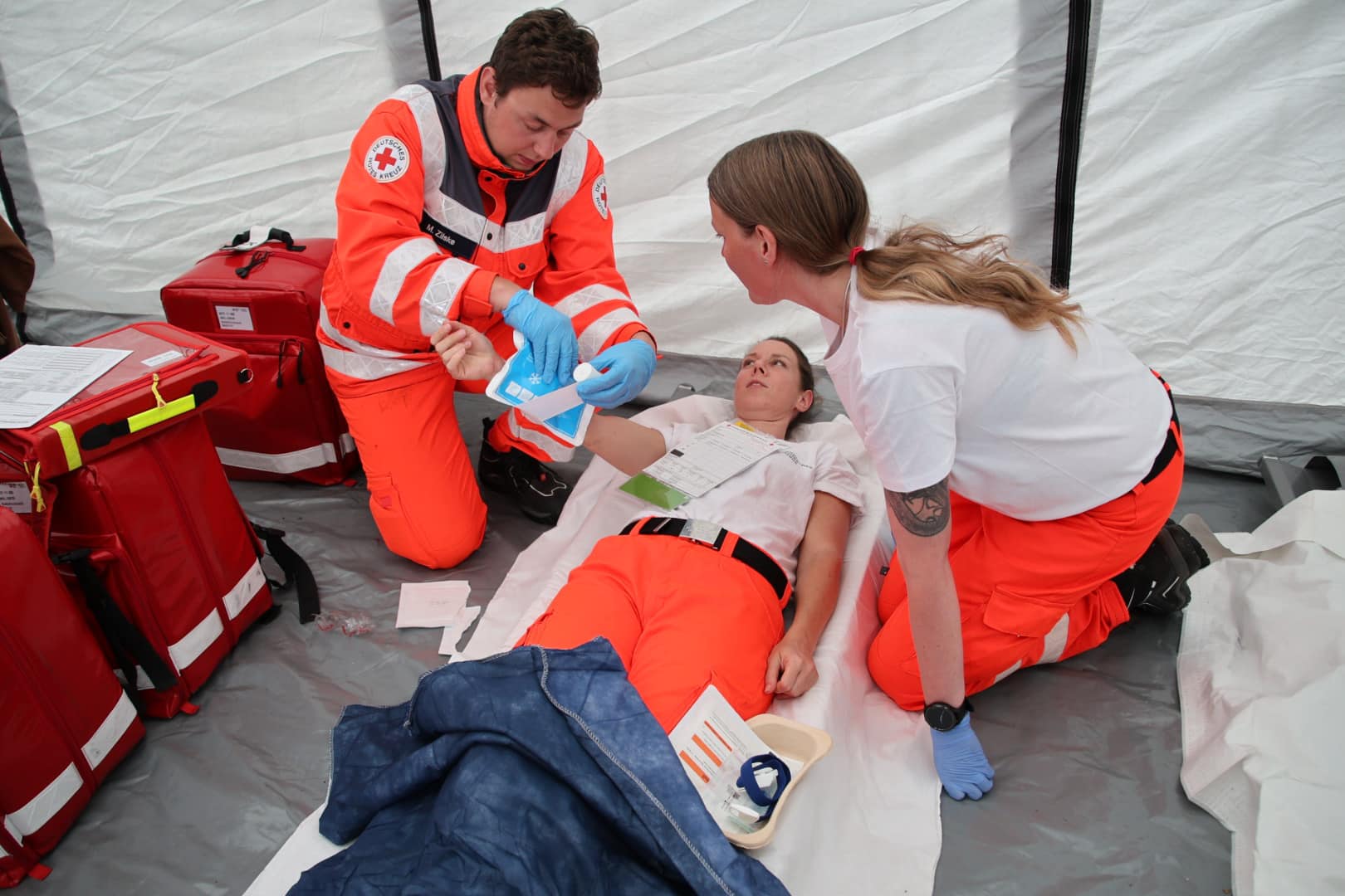 Sanitäter versorgen Person, die auf einer Trage liegt, in einem Zelt. Zwei Notfallkoffer sind sichtbar.