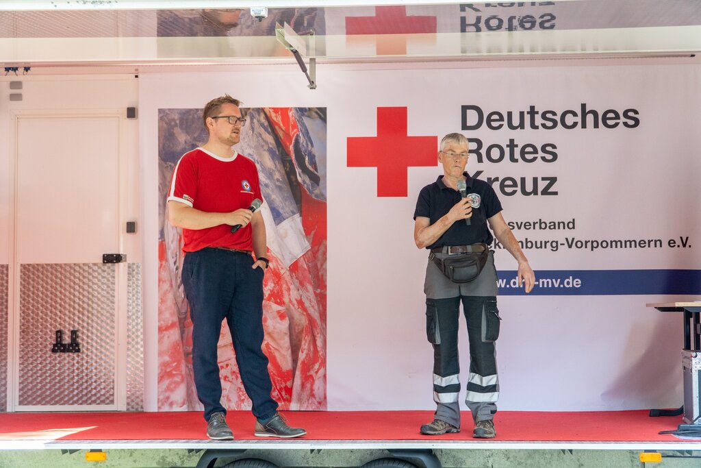 Zwei Männer mit Mikrofonen vor einem „Deutsches Rotes Kreuz“-Banner auf einer Bühne.