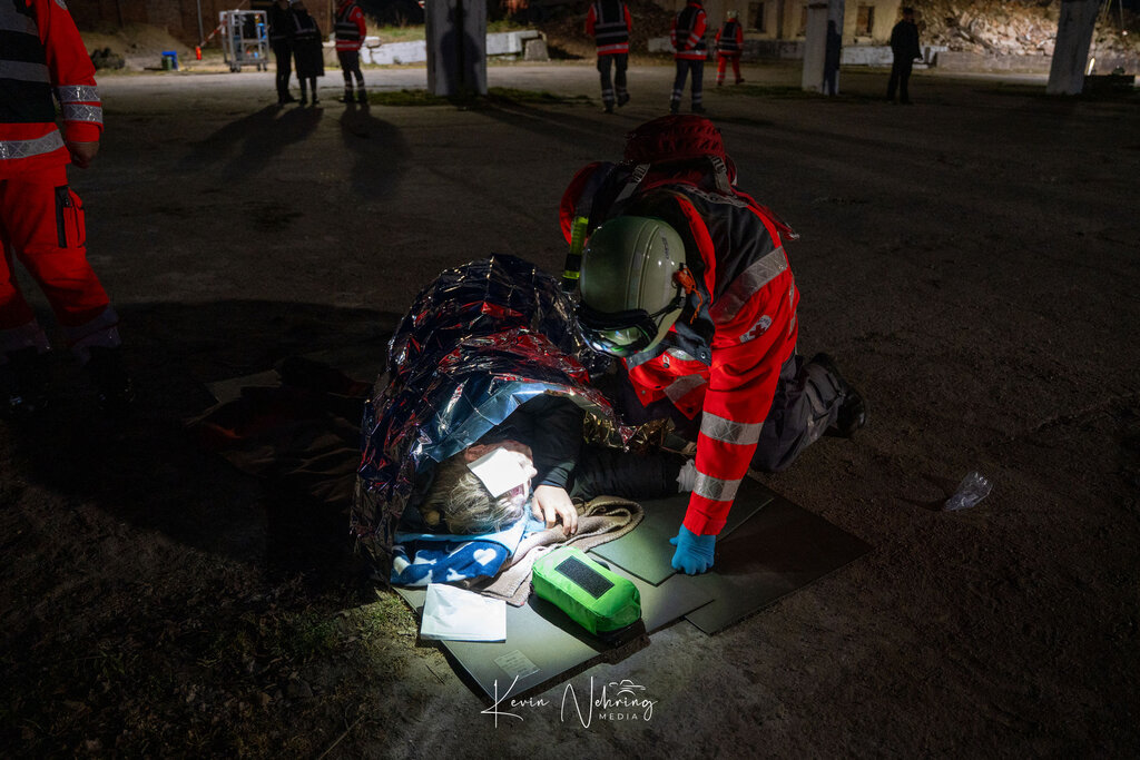 Sanitäter versorgt verletzte Person unter Rettungsdecke; Nacht, andere Helfer im Hintergrund.